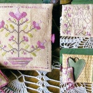 May include: A set of three needlepoint needle minders with a floral design. The needle minders are made of beige fabric with pink and green embroidery. The needle minders are in the shape of a square, a rectangle, and a heart.