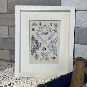 Op de afbeelding: Een wit houten frame met een witte passe-partout en een kruissteekontwerp erin. Het ontwerp is in tinten blauw en heeft een hart met het woord "Januari" erin. Het ontwerp is omgeven door verschillende geometrische vormen en bloemmotieven.