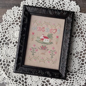 Könnte beinhalten: Ein gerahmtes Kreuzstich-Exemplar in einem schwarzen, verzierten Rahmen. Das Exemplar zeigt ein Haus, Blumen und die Buchstaben "ABC&J" in einem floralen Design. Die Farbpalette umfasst Beige, Rosa, Grün und Rot.