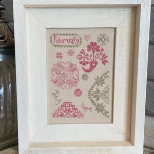 Op de afbeelding: Een ingelijst kruissteekkunstwerk met het woord "February" en diverse bloemen- en geometrische patronen in roze en groen. Het kunstwerk staat tegen een neutrale achtergrond en is ingelijst in een witte houten lijst.