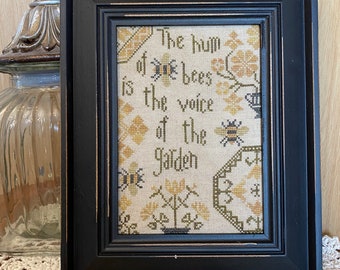 May include: A framed cross-stitch sampler with a black frame and a white background. The sampler features a quote about the sound of bees in a garden: "The hum of bees is the voice of the garden."