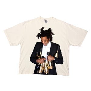 May include: An off-white short-sleeve t-shirt featuring a large graphic print of a person with dreadlocks wearing a black suit and white shirt, holding several gold-coloured champagne bottles. The shirt has a small tag at the neck reading WORLD FAMOUS.