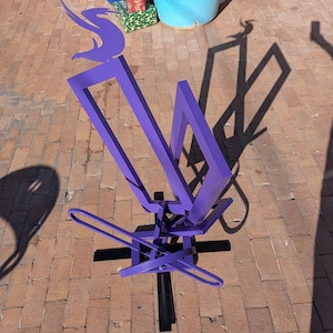 May include: A modern, abstract sculpture in a vibrant purple hue. The metal artwork features geometric shapes and curved lines, mounted on a black base. The sculpture's design creates interesting shadows on the brick surface.