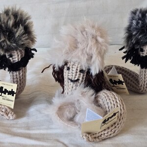 May include: Three handmade knitted dolls with furry hair and beards. Each doll has a beige body and a long, curved tail. They hold small cards with black text. The dolls are on a white surface.