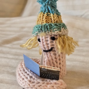 May include: A handmade knitted doll with a blue, yellow, and green hat, blonde yarn hair, and a pink body. The doll is reading a small blue book with the text "JOYCE CAROL BATES" visible. The doll is sitting on a white surface.