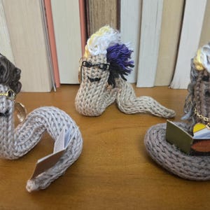 May include: Three whimsical, knitted worm figures with yarn hair, glasses, and jewelry. One worm holds a book. The worms are in shades of gray and brown, with colorful accents. They are displayed on a wooden surface.