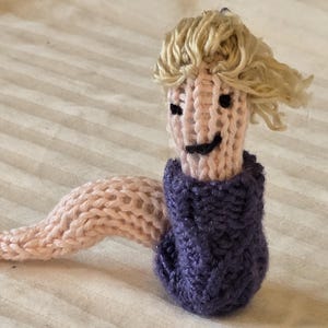以下が含まれることがあります： 手作りの編み物で作られた、笑顔の顔、金髪、紫色のセーターを着たワームのような生き物。ワームの体は薄いピンク色です。この編み物のおもちゃは、気まぐれでユニークなアイテムです。