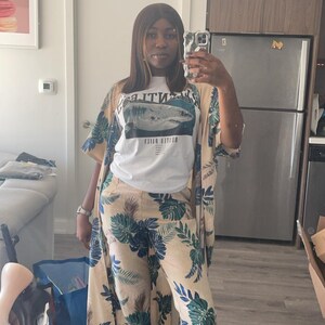 May include: A white t-shirt featuring a graphic of a shark and the text "HUSTLE DAILY" paired with a matching beige kimono and wide-leg trousers set, both decorated with a repeating pattern of dark blue and green tropical palm leaves.