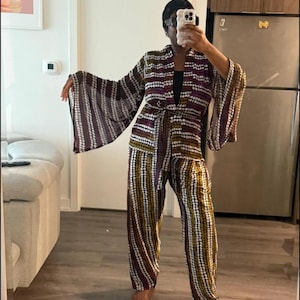 May include: A patterned jumpsuit with wide sleeves and matching pants. The jumpsuit features a belt and a design of vertical stripes in white, brown, and yellow. The outfit is worn in a home setting.