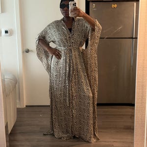May include: A full-length leopard print dress with wide sleeves and a belted waist. The dress is made of a lightweight fabric and has a flowing silhouette. The dress is worn with sunglasses.