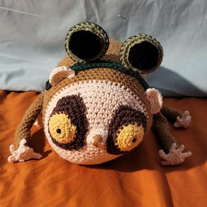 May include: A handmade crochet slow loris plush toy. The toy features a round head with large yellow eyes, a white and brown face, and small ears. It has brown arms and legs with white hands and feet. The toy has two large, round, green and black eye-like structures on its head.