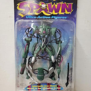 May include: A green and silver Spawn action figure in a plastic package. The figure is a cyborg with a robotic arm and a missile launcher. The package says "Cybernetic Arm Shoots Missile & has Whipping Action!"