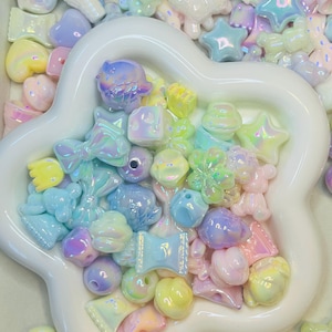 May include: A collection of iridescent pastel-colored beads in various shapes, including stars, bows, and hearts, are arranged in a white, flower-shaped dish. The beads have a glossy finish and reflect a rainbow of colors, creating a whimsical and playful aesthetic.