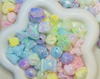 May include: A collection of iridescent pastel-colored beads in various shapes, including stars, bows, and hearts, are arranged in a white, flower-shaped dish. The beads have a glossy finish and reflect a rainbow of colors, creating a whimsical and playful aesthetic.