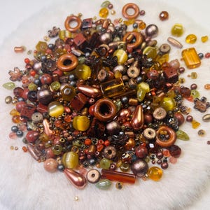 May include: A selection of mixed beads in diverse forms and dimensions. The beads display hues of brown, gold, green, and copper. Some are spherical, others are rectangular, and some are teardrop-shaped. The beads are arranged on a white, fluffy surface.