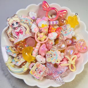 May include: A collection of assorted plastic charms and cabochons in a white scalloped bowl. The items feature anime-style characters with blonde hair, pink bows, yellow crescent moons, stars, ice cream cones, and iridescent butterfly shapes.