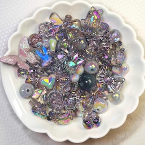 Puede incluir: Un plato blanco festoneado lleno de una variedad de cuentas y abalorios iridiscentes. La colección incluye formas de mariposas, corazones y conejos, junto con varias cuentas redondas e irregulares. Las cuentas tienen un efecto brillante y multicolor, que refleja la luz.