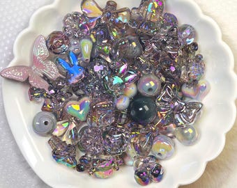 May include: A white scalloped dish filled with an assortment of iridescent beads and charms. The collection includes butterfly, heart, and bunny shapes, along with various round and irregular beads. The beads have a shimmering, multi-colored effect, reflecting light.