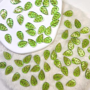 Può includere: Una collezione di perline in vetro traslucido a forma di foglia verde, ognuna caratterizzata da un dettagliato motivo a venature color oro. Questi piccoli componenti a forma di goccia sono sparsi su una superficie bianca e un materiale bianco testurizzato, mostrando un design botanico coerente.