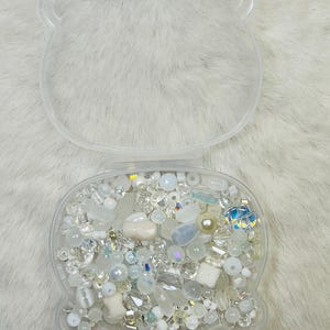 May include: A clear, bear-shaped container filled with an assortment of white and iridescent beads, crystals, and pearls. The container is open, revealing the contents, and sits on a white, fluffy surface. The beads vary in size and shape, creating a sparkling effect.
