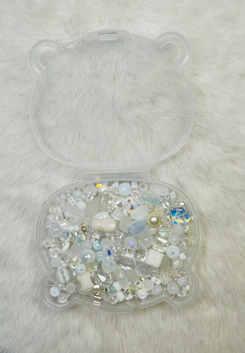 May include: A clear, bear-shaped container filled with an assortment of white and iridescent beads, crystals, and pearls. The container is open, revealing the contents, and sits on a white, fluffy surface. The beads vary in size and shape, creating a sparkling effect.