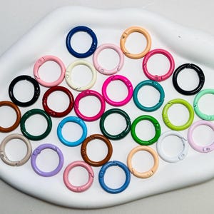 Könnte beinhalten: Eine Sammlung farbenfroher, runder Metall-Federringverschlüsse in verschiedenen Farbtönen, darunter Blau, Rosa, Grün und Braun. Die Ringe sind auf einem weißen, organisch geformten Tablett angeordnet. Diese Verschlüsse eignen sich ideal zum Basteln und für die Schmuckherstellung.