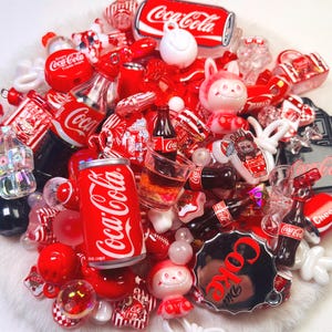 May include: A collection of miniature Coca-Cola themed charms and beads in red, white, and black. The assortment includes tiny soda cans, glass bottles, character figures, and circular bottle cap designs, all featuring the Coca-Cola brand logo.