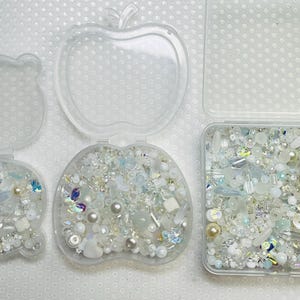 May include: Three clear plastic containers, shaped like a bear, an apple, and a square, each filled with an assortment of white, iridescent, and pale blue beads, pearls, and decorative elements. The containers are open, displaying the contents.