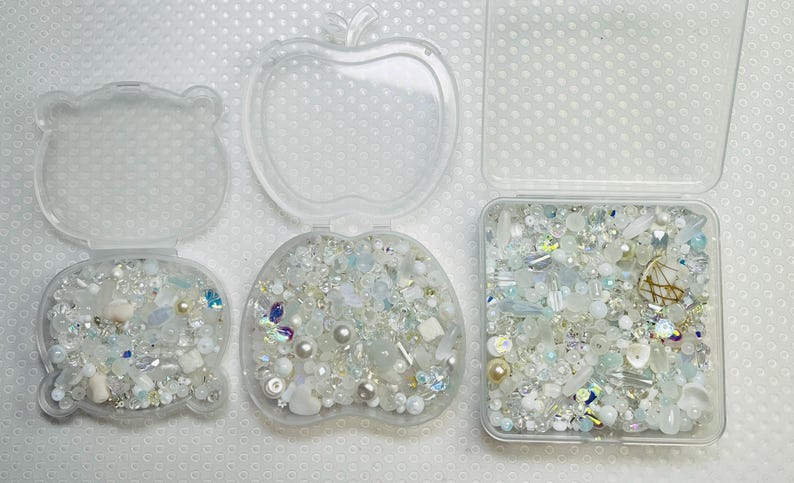 May include: Three clear plastic containers, shaped like a bear, an apple, and a square, each filled with an assortment of white, iridescent, and pale blue beads, pearls, and decorative elements. The containers are open, displaying the contents.