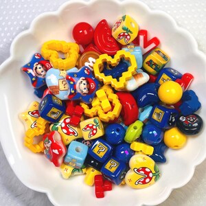 May include: A white scalloped bowl filled with colorful beads. The beads feature various shapes and designs, including stars, hearts, and cubes, with images of a popular video game character. The color palette includes red, yellow, blue, and green.