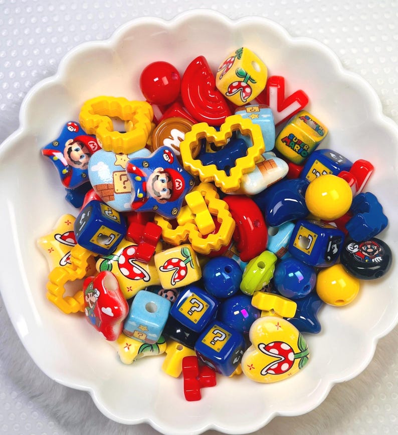 May include: A white scalloped bowl filled with colorful beads. The beads feature various shapes and designs, including stars, hearts, and cubes, with images of a popular video game character. The color palette includes red, yellow, blue, and green.