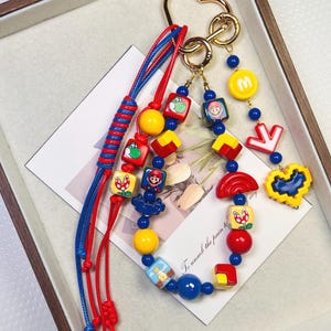 May include: A colorful keychain featuring various cartoon character charms. The keychain includes red and blue cords, gold-colored hardware, and a variety of charms in yellow, red, and blue. The charms depict characters and symbols.