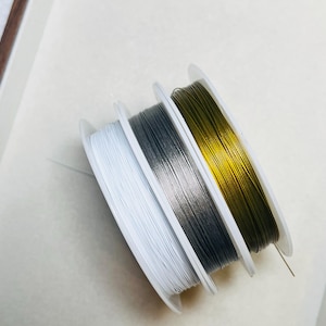 Puede incluir: Tres carretes de alambre en blanco, gris y dorado. El alambre está enrollado firmemente en carretes de plástico blanco. Los carretes están dispuestos en fila, con el carrete dorado a la derecha. El fondo es de color beige claro.