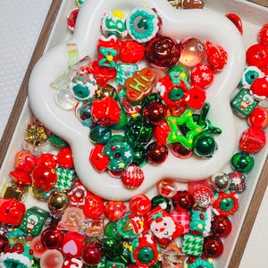 May include: A collection of Christmas-themed beads in a white, flower-shaped dish. The beads are red, green, gold, and white, with various shapes including round, square, and star. Some beads feature festive designs like snowmen and Christmas trees.