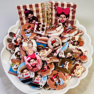 May include: A collection of small, flat, multi-shaped plastic charms featuring cartoon characters with brown fur and various outfits, including some with red bows, displayed in a white scalloped bowl behind a small spiral-bound notebook with a plaid cover.