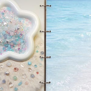 May include: A white star-shaped dish filled with a collection of small, translucent, and opaque beads in shades of light blue, pink, and clear. Additional loose beads are scattered on a white textured surface next to a page showing ocean waves.
