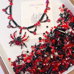 Op de afbeelding: Een handgemaakte ketting en spinnenhanger met rode en zwarte kralen. De ketting heeft zwarte buiskralen en rode accenten. Een kaart met de woorden "WELCOME TO" is zichtbaar. Een mix van rode, zwarte en heldere kralen vult het frame.