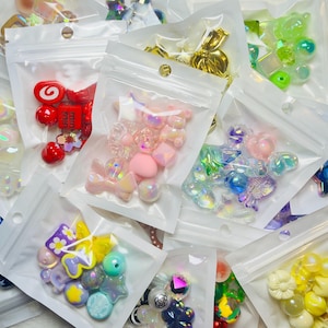 May include: A selection of small, colourful craft beads and embellishments in clear plastic zip bags. The assortment includes various shapes like bows, hearts, and geometric forms in shades of pink, yellow, green, and iridescent finishes. Ideal for jewellery making and crafting.