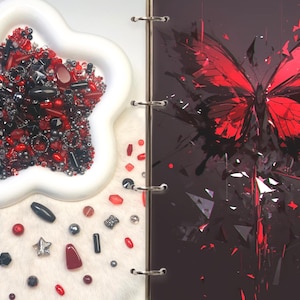 May include: A white flower-shaped tray filled with a collection of red, black, and silver beads and charms, positioned next to a dark binder page featuring a vibrant red and black shattered butterfly graphic with sharp geometric shards.
