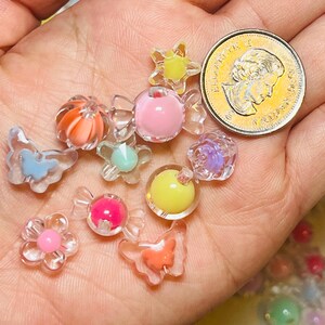 May include: A collection of small, colorful plastic beads in various shapes including butterflies, flowers, and candy wrappers, shown next to a Canadian one-dollar coin for scale. The beads feature translucent and opaque finishes in pink, yellow, orange, and blue.