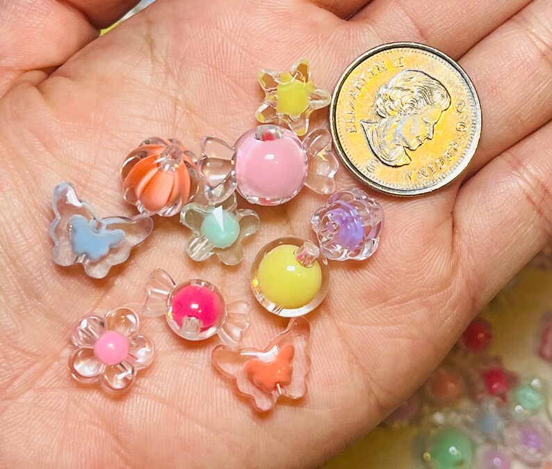 May include: A collection of small, colorful plastic beads in various shapes including butterflies, flowers, and candy wrappers, shown next to a Canadian one-dollar coin for scale. The beads feature translucent and opaque finishes in pink, yellow, orange, and blue.