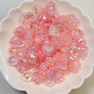Puede incluir: Un conjunto de abalorios de resina rosa iridiscente en diversas formas, incluyendo lazos, corazones, flores y formas geométricas. Los abalorios están dispuestos en un cuenco blanco con bordes festoneados. Estos elementos decorativos son ideales para manualidades y bisutería.