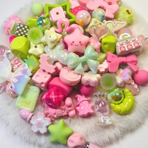 Può includere: Una collezione di perline assortite per il fai da te nei toni del rosa e del verde lime, con forme come stelle, fiocchi, cuori, fiori e una piccola borsa. Alcune perline hanno finiture iridescenti, mentre altre sono in plastica solida o traslucida.