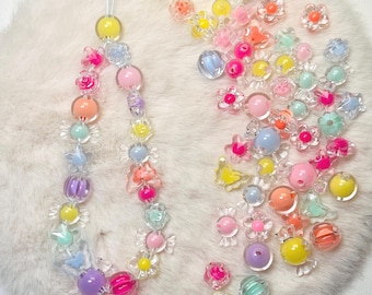 May include: A colorful beaded phone charm with a variety of pastel-colored beads, including flowers, stars, and round beads. The charm is strung on a clear cord and is surrounded by loose beads.