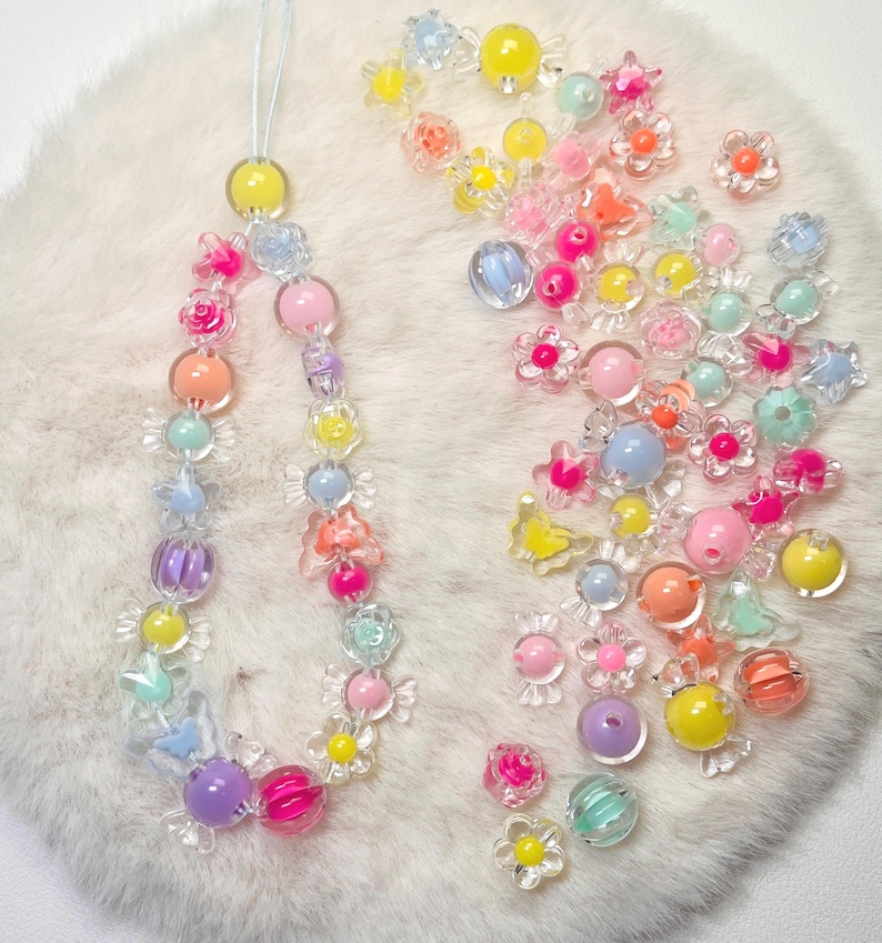 May include: A colorful beaded phone charm with a variety of pastel-colored beads, including flowers, stars, and round beads. The charm is strung on a clear cord and is surrounded by loose beads.