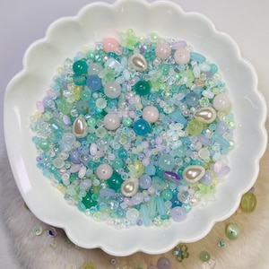 May include: A white, scalloped dish overflowing with beads in pastel shades of blue, green, pink, and white. The beads are diverse, including round, teardrop, and floral shapes, some with a pearlescent sheen. Ideal for jewellery making or other crafts.