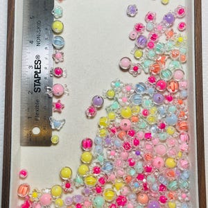 May include: A collection of colorful, translucent beads and embellishments in a wooden-framed tray. The assortment includes various shapes like flowers, spheres, and butterflies, in shades of pink, yellow, blue, and orange. A ruler shows measurements in inches.