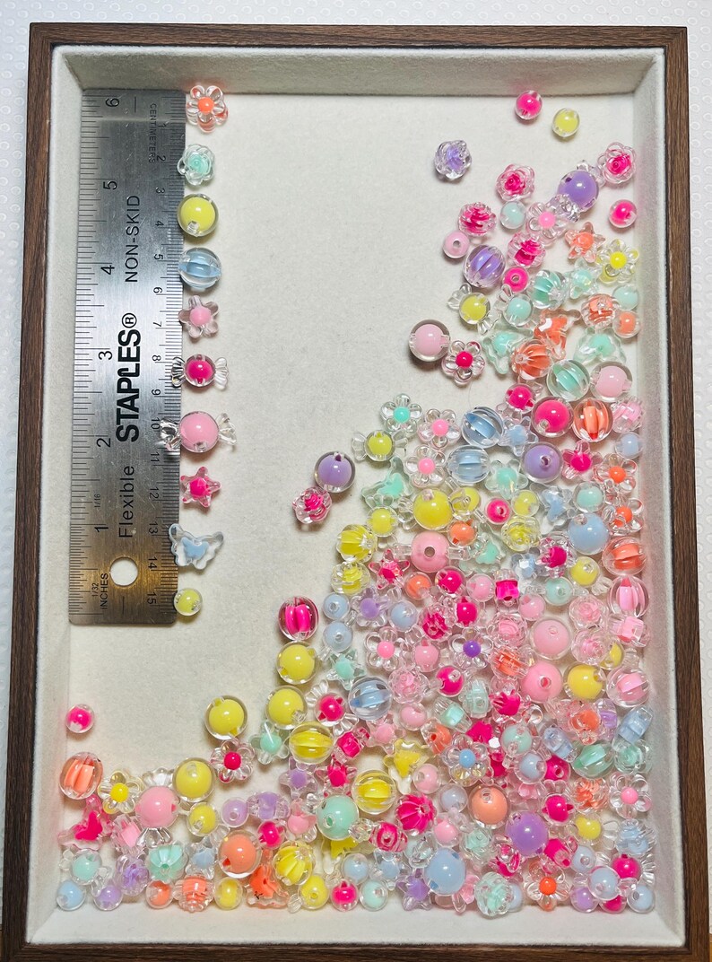 May include: A collection of colorful, translucent beads and embellishments in a wooden-framed tray. The assortment includes various shapes like flowers, spheres, and butterflies, in shades of pink, yellow, blue, and orange. A ruler shows measurements in inches.