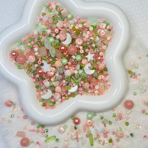 May include: A white, star-shaped dish filled with an assortment of beads in shades of pink, green, and peach. The beads include pearls, stars, moons, and various shapes, creating a colorful and textured display. The dish sits on a white, fluffy surface.