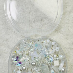 May include: Clear, apple-shaped container filled with an assortment of white and iridescent beads, pearls, and decorative elements. The beads vary in size and shape, including round, faceted, and floral designs. The container is open, revealing the contents.